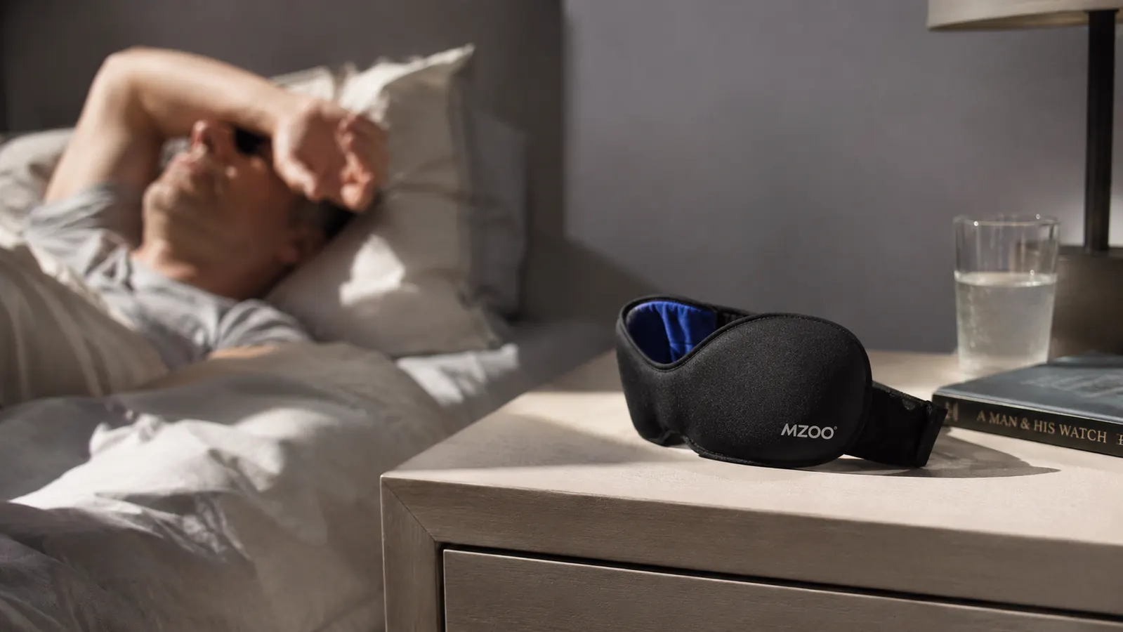 MZOO sleep mask lying unstrapped on a nightstand next to a lamp, afternoon light coming through a window behind it